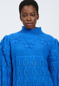 Maglione lavorato a maglia blu con colletto alto, motivi floreali testurizzati e corpo a fantasia con dettagli in pizzo.