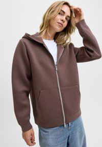 Felpa marrone con zip e cappuccio, due tasche frontali e polsini a costine. Indossata sopra una maglietta bianca, abbinata a jeans azzurri chiari. Tessuto morbido.