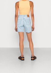 Světlé džíny šortky s roztrhaným lemem, zadními kapsami a koženou značkovou nášivkou. Spárováno s přiléhavým oranžovým tílkem a tmavými sandály.