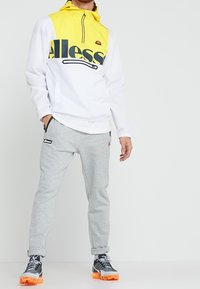 White and yellow hooded windbreaker with large black logo, paired with grey sweatpants and black sneakers with orange accents.