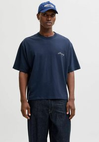 Marineblå bomulls-T-skjorte med avslappet passform, korte ermer og brodert logo på brystet. Kombinert med mørke denimbukser.