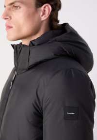 Svart pufferjakke med høy krage og hette. Har et firkantet logo-patch på venstre arm. Glatt, shiny stoff med et elegant design.