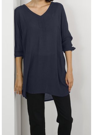 Marineblaue Tunika mit V-Ausschnitt und dreiviertellangen, hochgerollten Ärmeln, im entspannten Schnitt und aus weichem, strukturiertem Stoff. Kombiniert mit schwarzen Hosen.