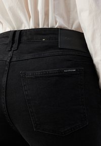 Gros plan d'une personne portant un jean en denim noir Marc O'Polo avec une poche arrière visible et une étiquette de marque, associé à une chemise blanche rentrée.