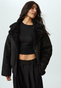 Crna jakna s umjetnim krznom i ovratnikom, koja se odlikuje mekim teksturama i zatvaranjem na gumbi. Uparena s crnim skraćenim topom i krojenim hlačama.