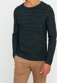 Mörkgrön långärmad stickad skjorta med rund halsringning och ett texturerat randigt mönster. Matchad med ljusbruna byxor.