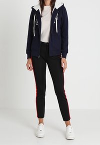 Marineblaue Zip-Up-Hoodie mit weißem Fleece-Innenfutter, seitlichen Taschen und Kordelzug; kombiniert mit schwarzen Hosen mit roten Seitenstreifen und weißen Sneakern.