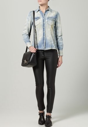 Camicia di jeans blu chiaro con due tasche sul petto, abbinata a pantaloni skinny in pelle nera e stivaletti alla caviglia. La modella tiene una borsa nera strutturata.