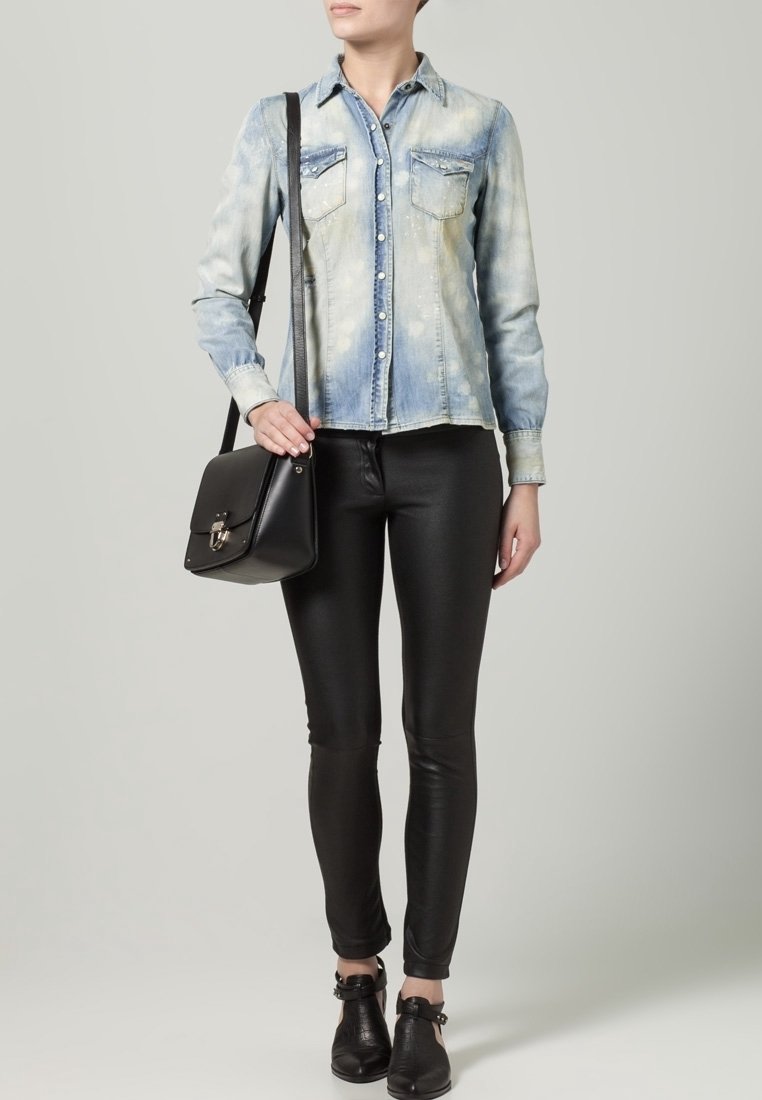 Camicia di jeans blu chiaro con due tasche sul petto, abbinata a pantaloni skinny in pelle nera e stivaletti alla caviglia. La modella tiene una borsa nera strutturata.