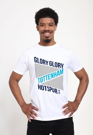 Mężczyzna uśmiechający się, stojący z rękami na biodrach, ubrany w białą koszulkę z napisem "Glory Glory Tottenham Hotspur" oraz logo drużyny na jednolitym tle.