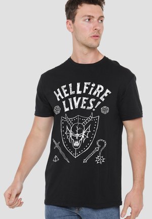 Young man wearing black T-shirt with white "HELLFIRE LIVES!" text, skull with horns on shield, two dice, sword, and mace illustrations.