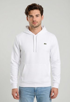 Giovane uomo con felpa bianca con cappuccio e tasca frontale, con piccolo logo di coccodrillo verde, abbinata a jeans azzurri.