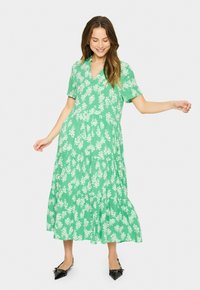 Rochie florală verde cu mâneci scurte, fustă în straturi și decolteu în v. Prezintă un model cu flori albe și un material ușor. Pantofi negri cu vârf ascuțit.