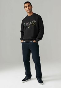Sudadera negra con gráficos coloridos de temática navideña, presenta un cuello redondo y puños de canalé, combinada con pantalones oscuros y zapatillas.