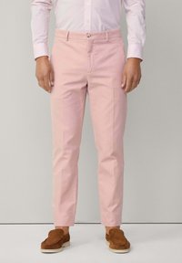 Pantaloni di cotone rosa con un design aderente, fronte piatto e tasche laterali. Abbinati a scarpe marroni slip-on. Camicia a righe bianca e rosa.