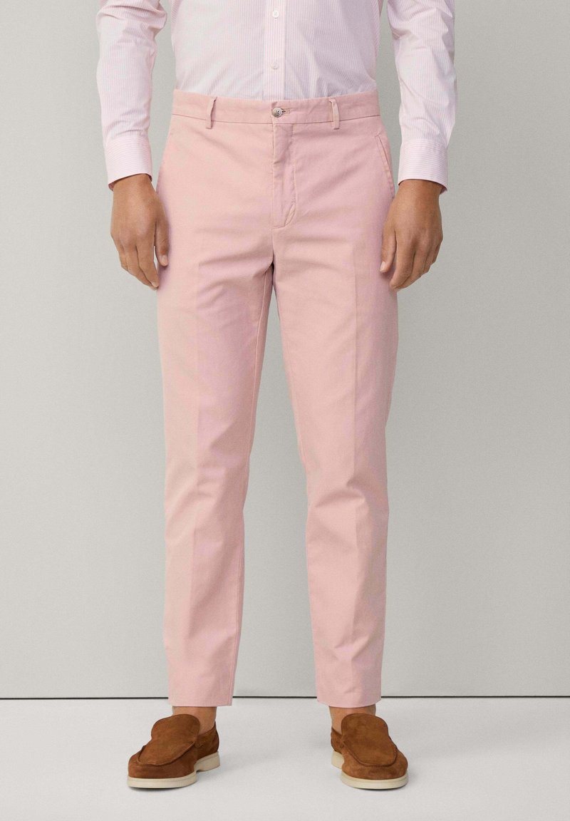 Pantaloni di cotone rosa con un design aderente, fronte piatto e tasche laterali. Abbinati a scarpe marroni slip-on. Camicia a righe bianca e rosa.