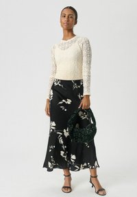 Vrouw in een crèmekleurige kanten top met lange mouwen, zwarte bloemen midi-jurk, zwarte haksandalen, houdend een textuur donkergroene handtas.