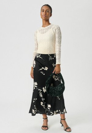 Woman wearing a cream lace long-sleeve top, black floral midi skirt, black heeled sandals, holding a textured dark green handbag.