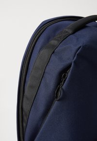 Zaino blu navy con tessuto texturizzato, dotato di una tasca con zip laterale e una cintura in nylon nera lungo la parte superiore per un facile trasporto.