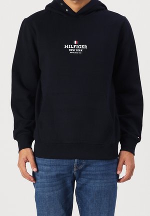 Uomo che indossa un felpa nera con il testo e il logo "HILFIGER NEW YORK" sul davanti, abbinata a jeans blu, in piedi con le braccia rilassate.