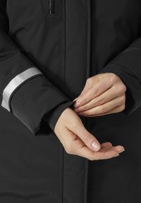 Veste noire imperméable avec une surface texturée. Présente une longue manche avec un accent réfléchissant argenté et un poignet ajusté. Mains partiellement visibles.