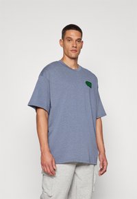 Blå bomulds t-shirt med korte ærmer og en afslappet pasform. Har et grønt logo-patch på venstre side. Komplementeret af grå sweats.