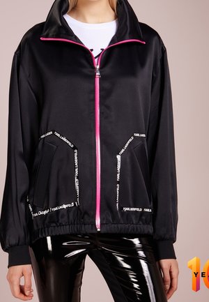 Zwarte jas met ritssluiting, roze rits en witte "KARL LAGERFELD" tekst rond de zakken, gecombineerd met glanzende zwarte broek.