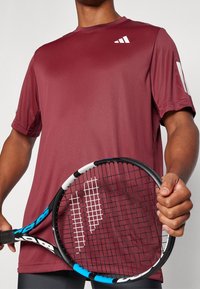 Camiseta atlética burdeos de manga corta, fabricada en un tejido suave, presenta un logo en el pecho y se acompaña de una raqueta de tenis.