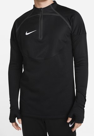 Mann trägt schwarzen Nike Langarm-Pullover mit Daumenlöchern und Viertelreißverschluss, kombiniert mit schwarzen Nike-Hosen mit weißen Swoosh-Logos.