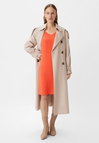Beige Wollmischung-Trenchcoat mit großen braunen Knöpfen, kombiniert mit einem knielangen orangefarbenen Kleid und beigen Knöchelstiefeln, auf einem neutralen Hintergrund.