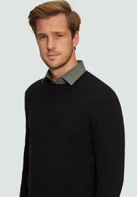 Schwarzer gestrickter Pullover mit Rundhalsausschnitt über einem grünen karierten Hemd mit Kragen. Enganliegender, strukturierter Stoff mit langen Ärmeln. Keine sichtbaren Muster.