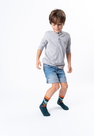 Young boy wearing gray long-sleeve shirt, rolled denim shorts, and colorful socks with dinosaur design dancing on a white background.