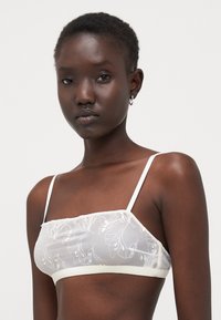 Hvit blonde-bikini med blomsterbroderi, gjennomsiktig stoff og tynne stropper. Har et mykt elastisk bånd nederst for komfort.