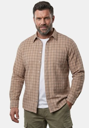 Mann mit kurzen dunklen Haaren und Bart, der ein hellbraunes kariertes Hemd über einem weißen T-Shirt und olivgrünen Cargohosen trägt.