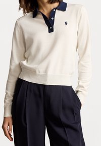 Pull en tricot crème avec un col marine, arborant un petit logo sur la poitrine. Le design comprend des poignets et un ourlet côtelés pour un meilleur ajustement.