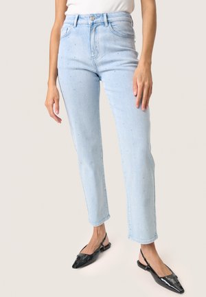 Femeie purtând blugi de talie înaltă, bleu deschis, cu mici detalii de perle, top alb și pantofi slingback negri, cu vârf ascuțit.
