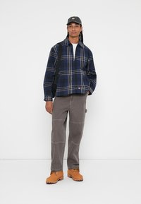Casaco de flanela com padrão xadrez azul-marinho e cinza com fecho, calças cargo cinzas com costura de destaque, e sapatos duráveis em tom de camelo. Modelo em pé segurando uma mala de ombro.