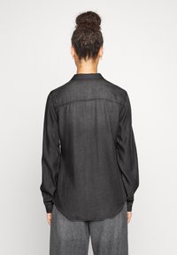 Camicia di denim nera con maniche lunghe, colletto classico e orlo curvo. Presenta cuciture visibili e una texture liscia. Design minimalista.