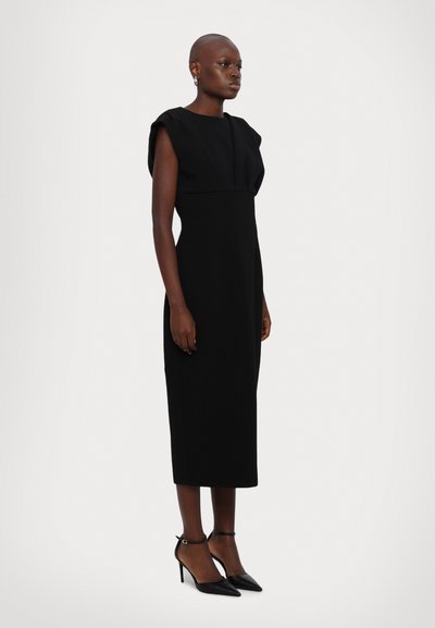 Black fitted midi dress with structured puff sleeves and a pleated bodice. Worn with black stiletto heels and ankle strap detail.
