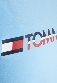 Světle modré bavlněné tričko s výrazným potiskem "TOMMY" v černé, červené a bílé barvě, s geometrickým logem umístěným vlevo.