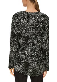 Zwarte blouse met lange mouwen en een gespikkeld wit patroon. Ronde halslijn en knoopschoenen, met een gebogen zoom en een relaxte pasvorm.