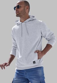 Haut à capuche gris clair avec cordon de serrage, poche latérale et poignets côtelés. Porté avec un short cargo bleu, le modèle affiche une coupe décontractée.