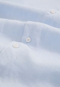 Zbliżenie jasnoniebieskiej lnianej tkaniny z białymi guzikami i widocznym przeszyciem na kołnierzyku i mankiecie koszuli.