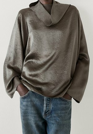 Personne portant un haut ample et texturé vert olive à manches longues avec un large col et un jean en denim bleu, mains dans les poches.