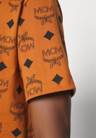 Orange bomullströja med svart MCM-logotryck, med geometriska mönster och rundad halsringning. Korta ärmar avslutar designen.