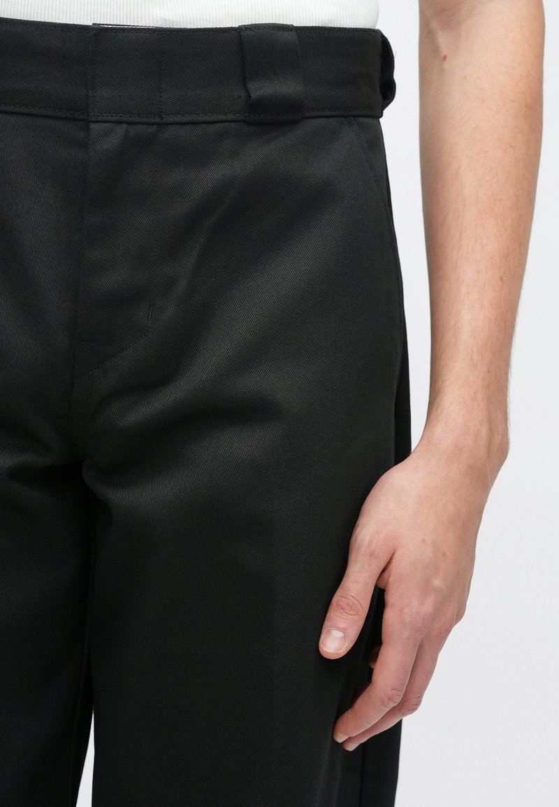 Sorte skræddersyede bukser med flad front, bæltestropper og en glat tekstur, hvor en hånd hviler på sømmen.