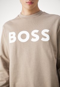 Sudadera beige de mezcla de algodón, con cuello redondo y gran logo blanco "BOSS" en el frente. Textura suave, corte relajado.