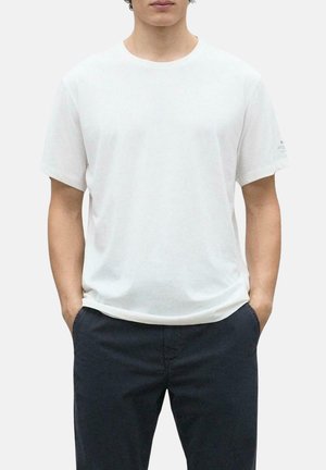 Camiseta de algodón blanca de cuello redondo con mangas cortas, corte relajado, textura suave y sutil detalle del logotipo en la manga izquierda. Llevada con pantalones oscuros.