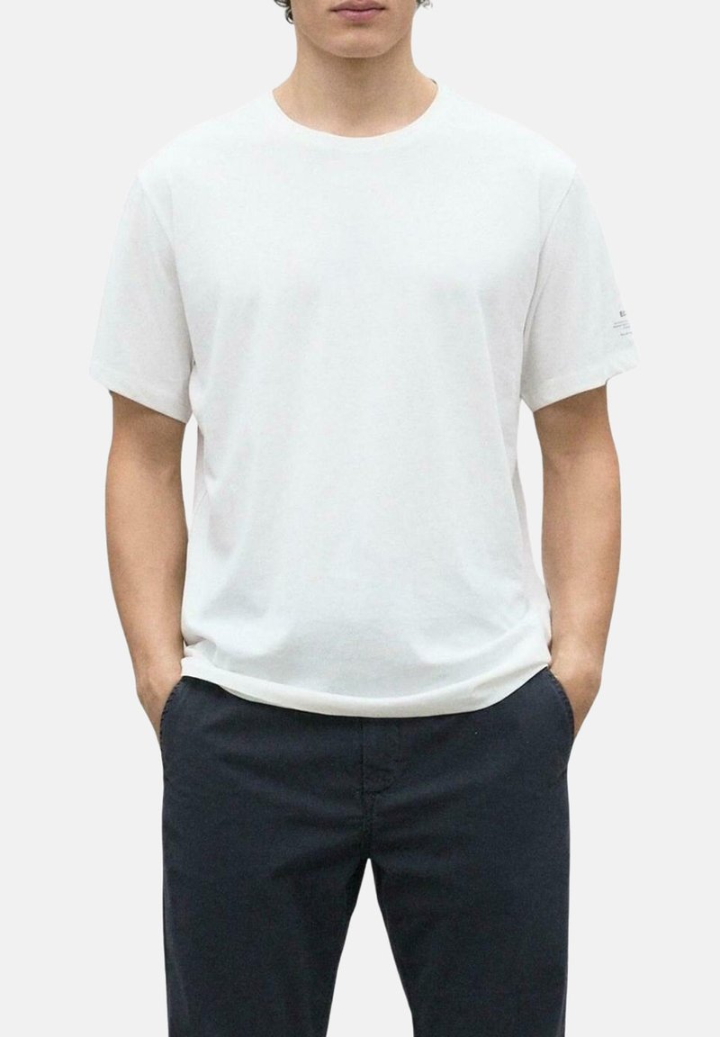 Camiseta de algodón blanca de cuello redondo con mangas cortas, corte relajado, textura suave y sutil detalle del logotipo en la manga izquierda. Llevada con pantalones oscuros.