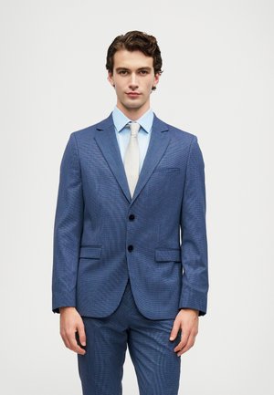 Young man wearing a blue checked suit, light blue shirt, and light beige tie, standing against a plain white background.